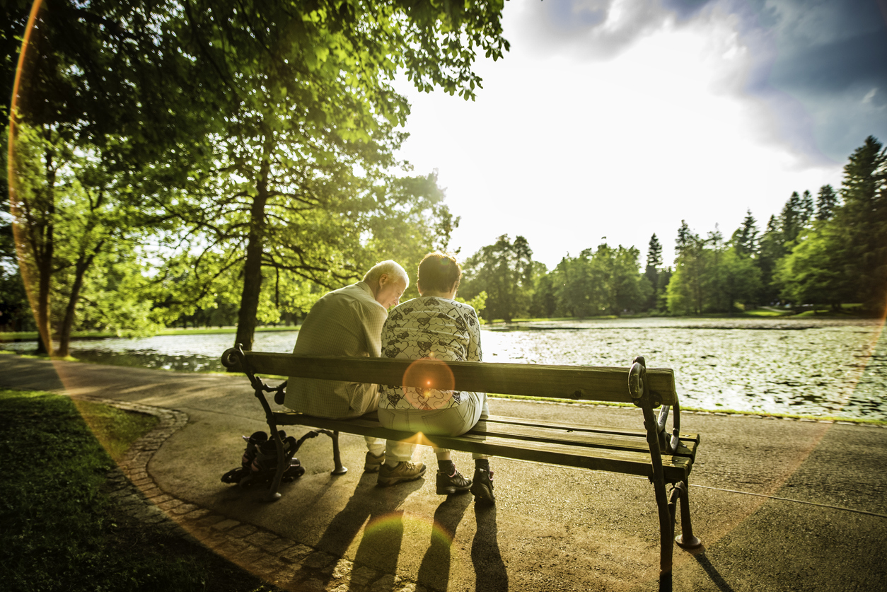 Rear view of a senior couple sitting on a bench near a lake in a park on a sunny day.