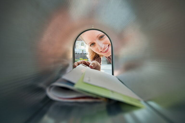 A woman grabbing mail from her mailbox. We are looking at her as if we are inside the mailbox.