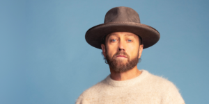 TobyMac in a brown hat in front of a sky-blue background. He is wearing a beige sweater.